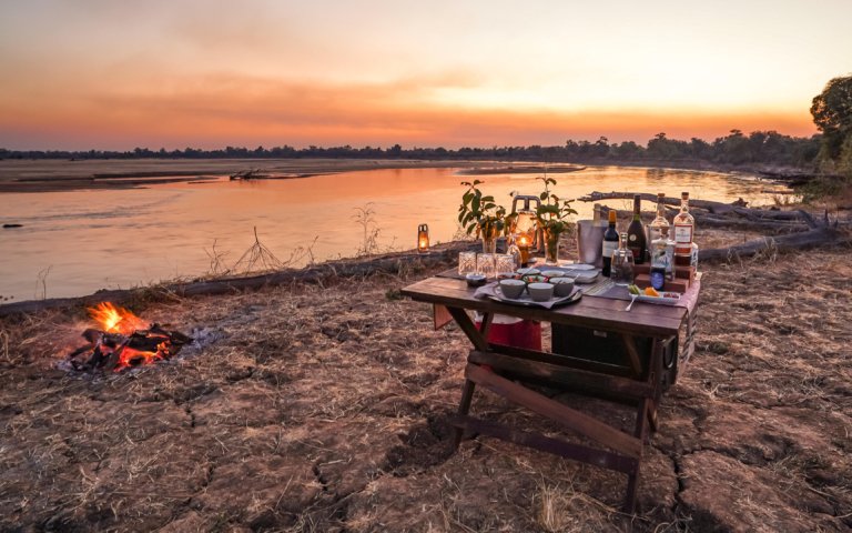 Chikunto Safari Lodge Sundower Tisch mit Getränken am Flussufer bei Dämmerung